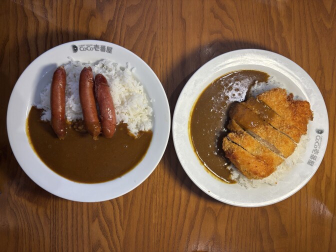 Two white plates sit against a brown wooden surface. The plate on the left contains a brown sauce with white rice and three cooked sausages on top. Next to it, the plate on the right contains the same brown sauce and white rice, with a light brown fried cutlet on top.  