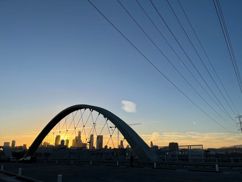 The skyline of downtown Los Angeles is visible at sunset behind one of the arches of the 6th Street bridge.