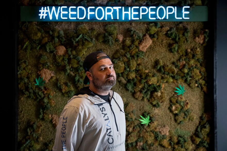 A white-looking man looks seriously at the camera. A sign behind him reads "#weedforthepeople". He wears a backwards baseball hat and athletic wear shirt.