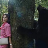 Keri Russell stands with her back against a large tree in a forest. She wears a bright pink shirt dress with a  white sling bag. Russell looks out the corners of her eyes, trying to see the other side of the tree without moving. She seems to be hiding. trying to stay as still as possible. On the other side of the tree is a large black bear sniffing the branches. It has its paws up against both sides of the tree, bracing itself on its two legs. 
