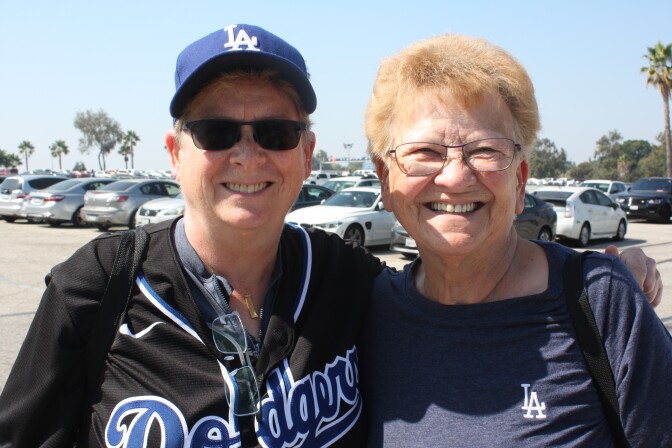 Two women in Dodger jerseys huddle together. They have short hair. The woman on the right is wearing glasses; the woman on the left is wearing sunglasses and a  Dodgers baseball hat. 