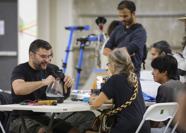 Two people sit at a table. One person with a beard is holding a coffee carafe; another with a long grey braid is sitting opposite them.