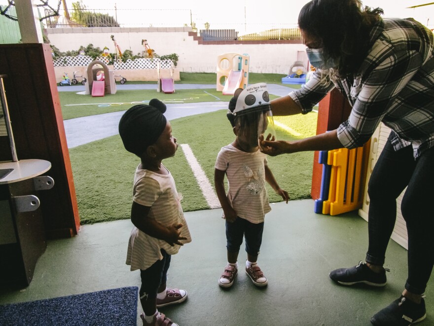 In this image, an adult helps a child put on a panda face shield, while another child watches.