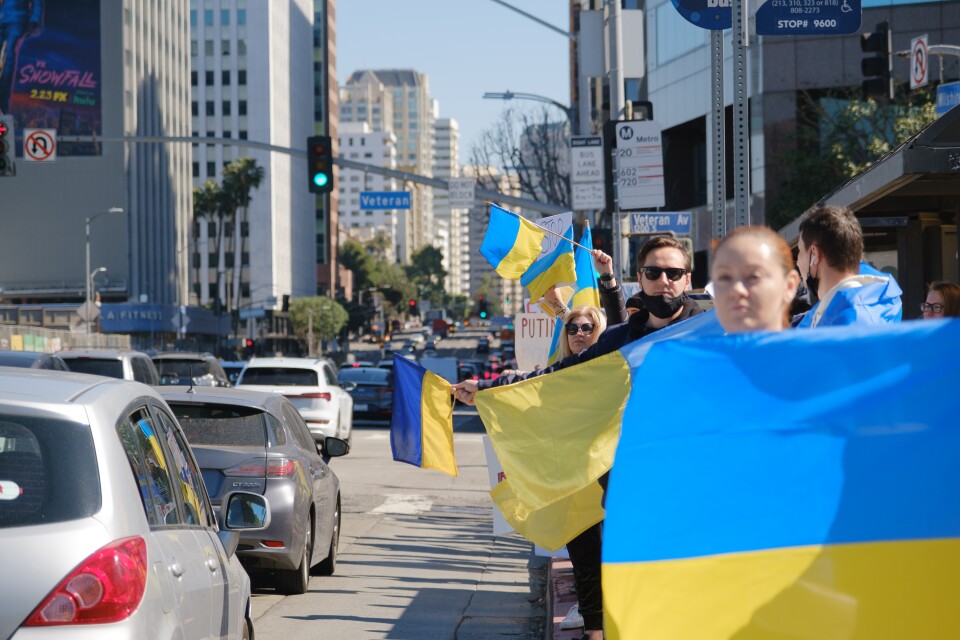 Several people stand on the side of a busy urban street holding Ukrainian flags.