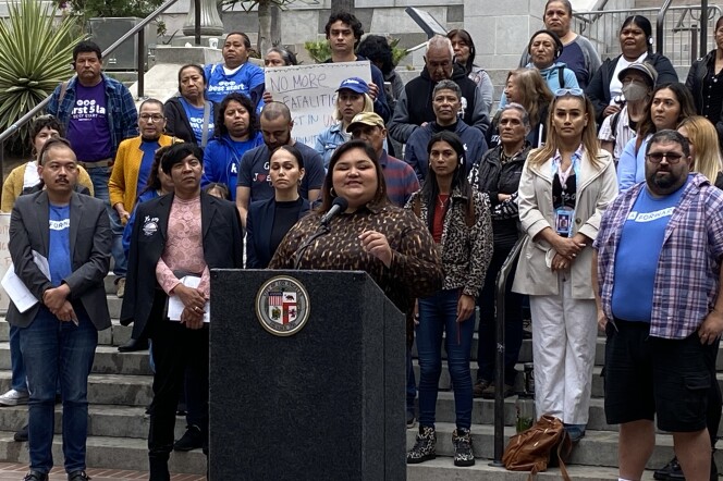 LA City Council member Eunisses Hernandez speaks at a podium that has the seal of Los Angeles on it. There is a crowd of more than a dozen behind her. Some wear blue t-shirts that mark them as supporters of the progressive policy advocacy group LA Forward. 
