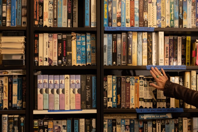 A hand reaches out towards a display wall full of VHS tapes. 