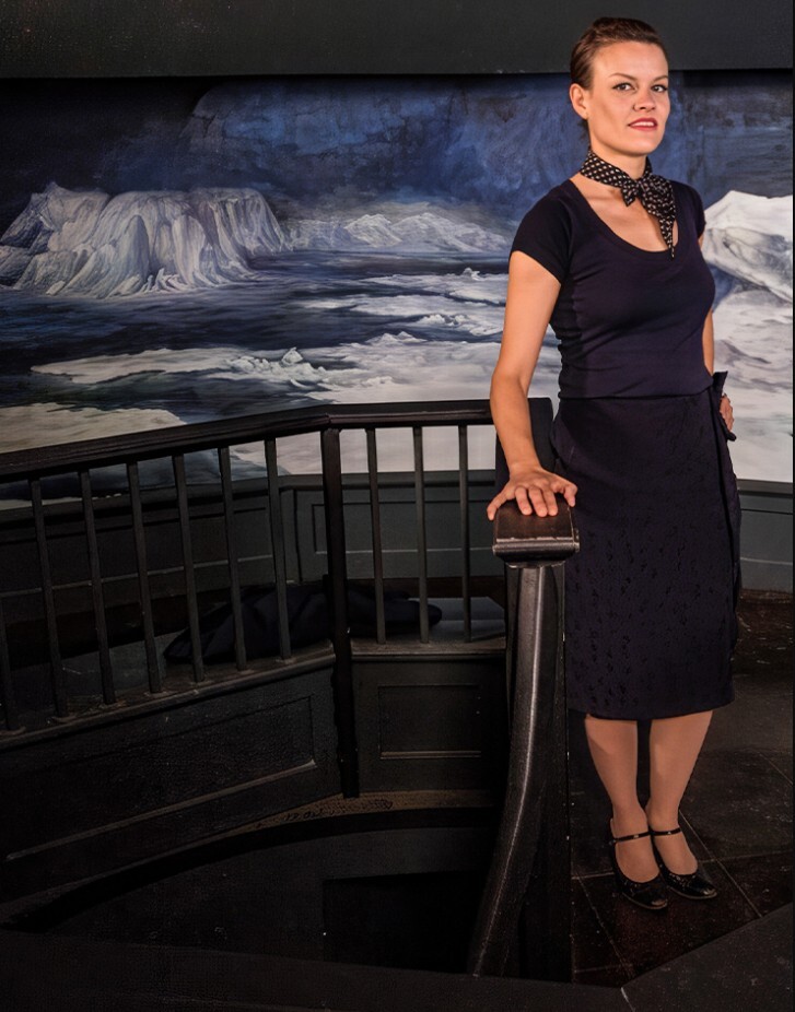 Woman with short dark hair and wearing a black dress poses before a painting.