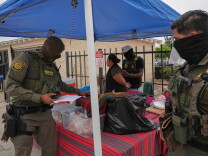 A woman standing underneath a blue pop up tent is detained by U.S. Border Patrol agents wearing army green uniforms. Underneath the tent are tables covered with red and grey striped tablecloths with various boxes and items.   