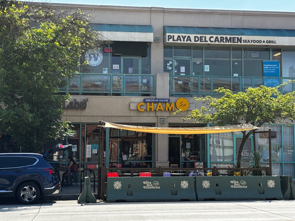 The exterior of Chamo, a Venezuelan restaurant in a Pasadena strip mall, with its bright yellow sign above the entrance and an outdoor dining area shaded by a canopy.