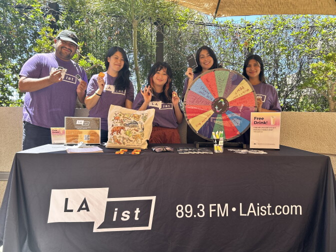 A group of five people hold small items and stand behind a table with a tablecloth that reads "LAist. 89.3 FM. LAist.com" with a spinning wheel and other items on top of it. 