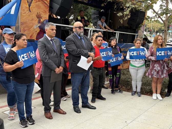 About a dozen people stand side-to-side on a public sidewalk. Many are golding signs that read: "ICE out of LA."