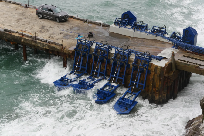 Blue paddle-like devices immersed in ocean surf are attached to a wooden pier. An SUV is parked on the pier.