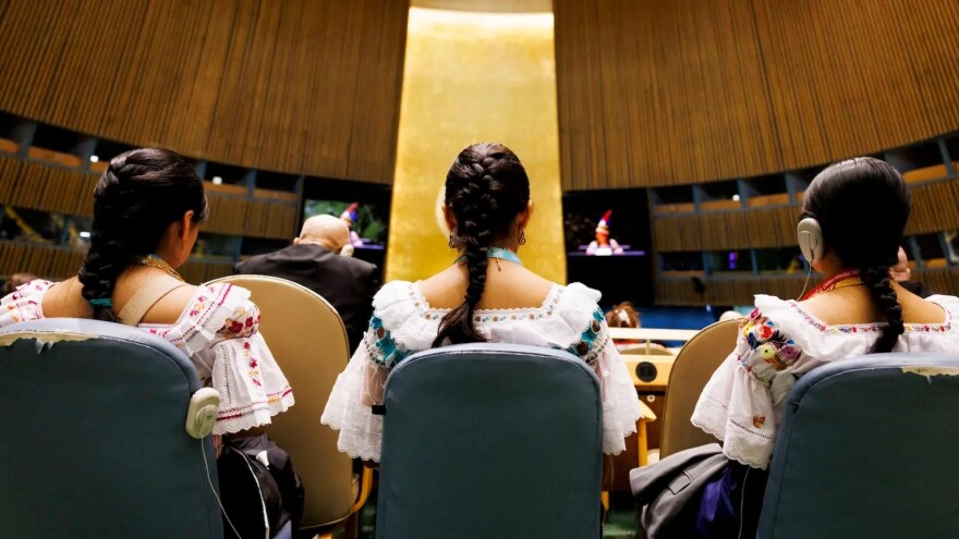 Three women are pictured from behind while sitting in blue chairs in a large, circular room. They each wear their hair in a single braid and are wearing white dresses with colorful embroidery.