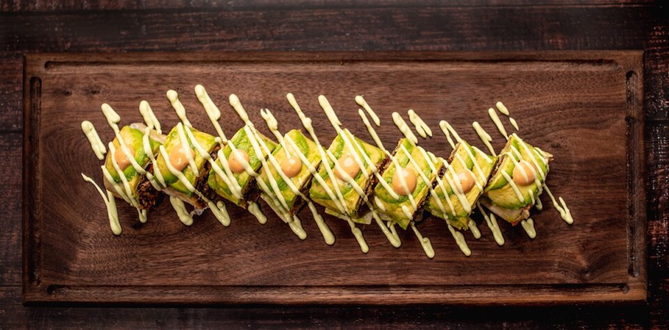 an overhead shot of a bunch of sushi rolls topped with avocado and drizzles of a creamy white sauce, all on top of a wood cutting board