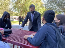 An adult wearing a suit leans over an outdoor table surrounded by middle school aged students.