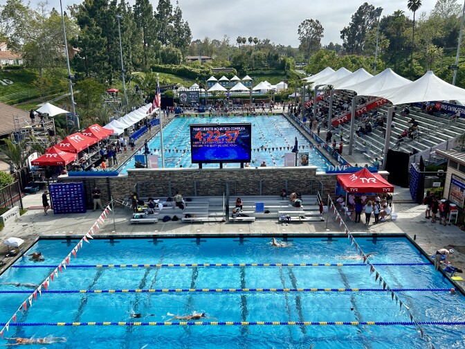 Looking down from above on two competition swimming pools. The one in the back has covered bleachers along the side and a large score board at the near end. 