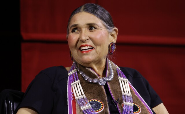 A woman has her hair pulled back, red lipstick and wears traditional Native American dress over her shirt.