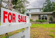 sign in foreground of photo in front of a house and overcast sky reads "FOR SALE" in all caps + red letters.