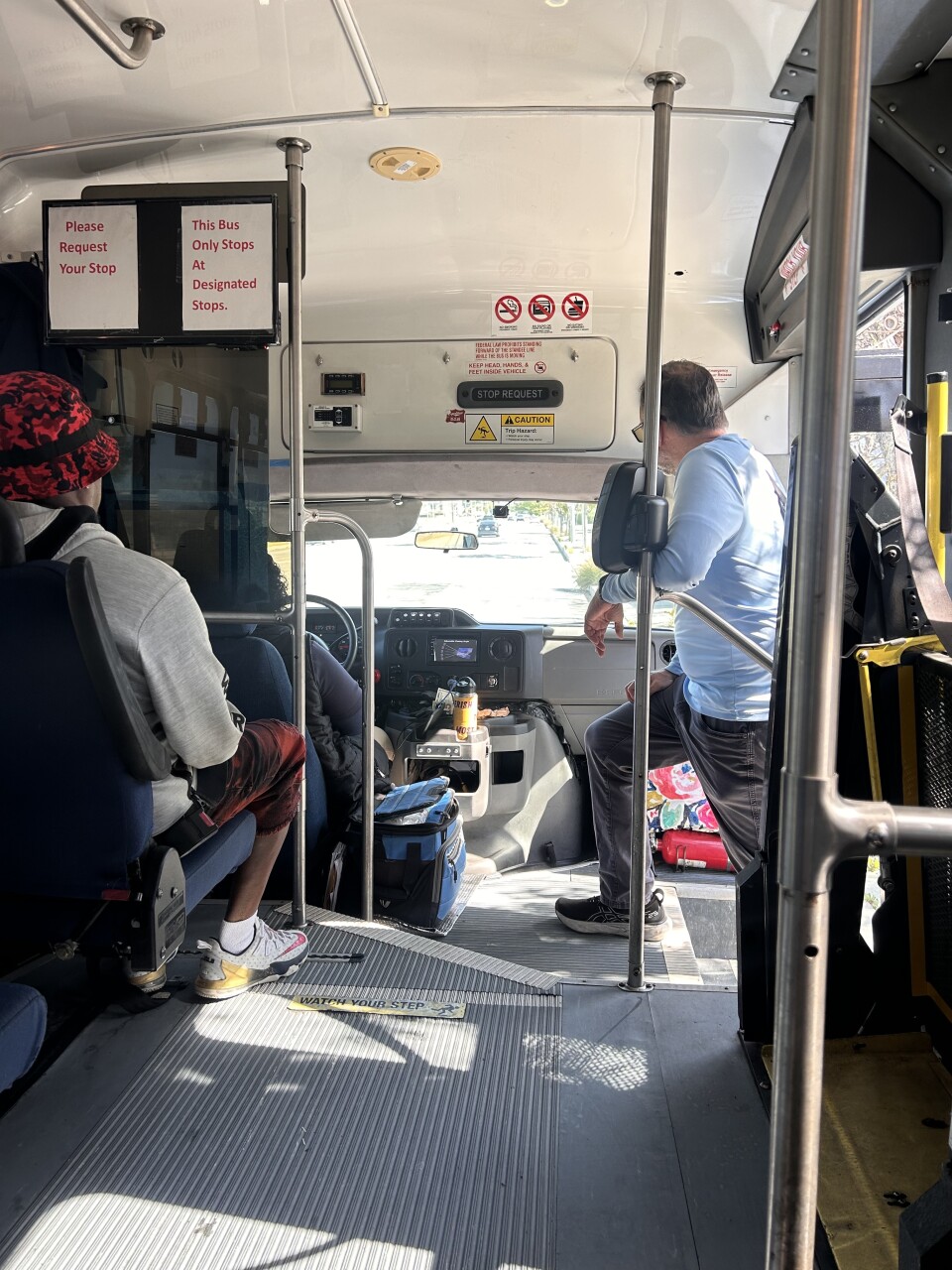 The interior of a small bus, with several silver metal poles protruding from the floor. A man is sitting in the first row of seats and another man is standing near the front entrance of the vehicle.