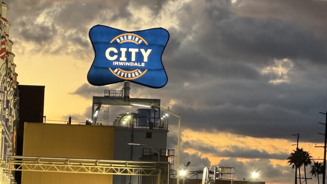 A big blue sign that say "City Brewing & Beverage Irwindale" is seen at sunset. Lights illuminate the sign. Large clouds can be seen behind it. Palm trees and power lines are seen in the right hand corner.