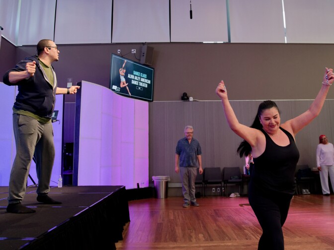 A man on a stage wearing a headset microphone and blue hoodie leads a dance class, while a woman in a black tank top and leggings dances in the foreground with her arms raised joyfully. A monitor in the background displays "DANCE CLASS ALVIN AILEY AMERICAN DANCE THEATER.