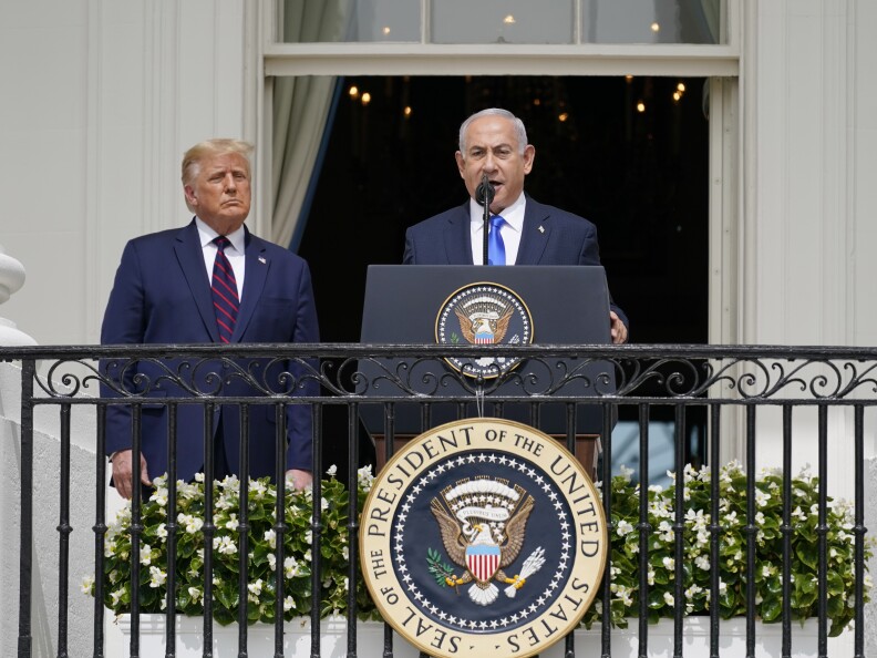 Two white me in suits are on a balcony with a wrought iron railing. on that railing is the seal of the President of the United States. One man speaks at a lectern.