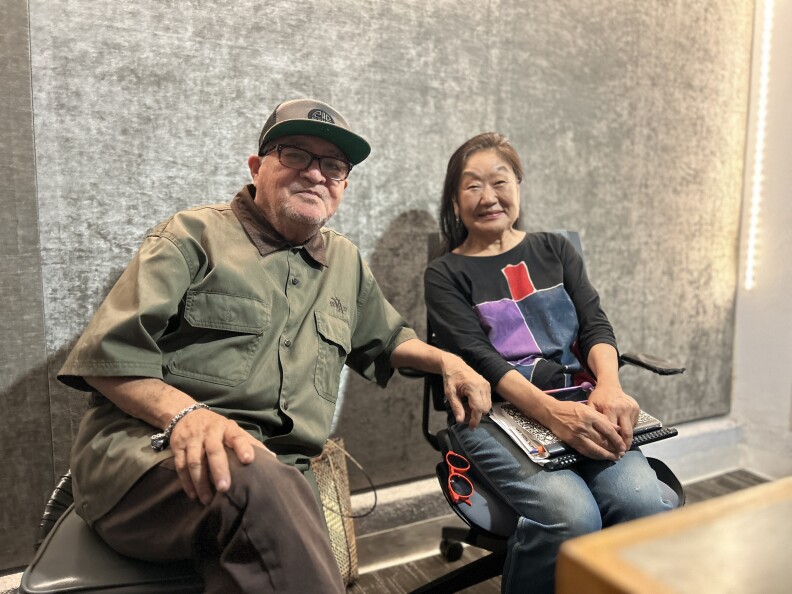 An older man with light skin man wearing a trucker hate and glasses sits in a chair beside a Japanese woman wearing a black quarter-sleeve shirt with color blocks and jeans. They smile for the camera in a radio studio with silver velvet walls. 