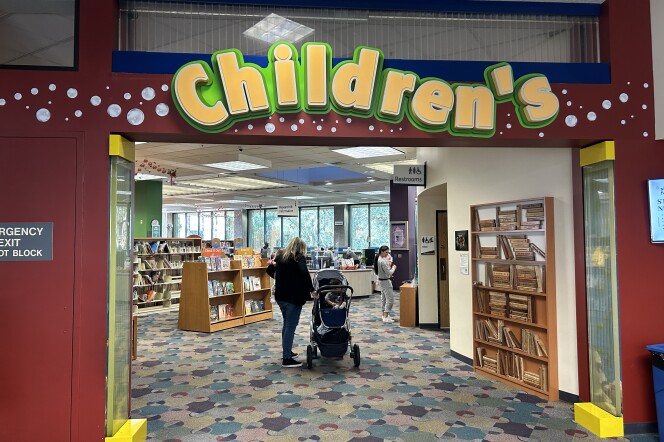 An entryway with a large yellow sign above that reads "Children's." Through the entry, there's a female-presenting person next to a baby stroller and shelves of books in the distance. 