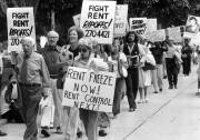A black and white view of a crowd of protesters walking up the sidewalk. The crowd is mostly older, white men and women who are carrying signs calling for a rent freeze and then rent control. One sign says to fight rent ripoffs.