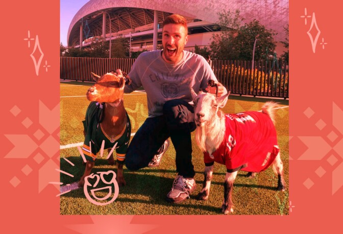 An image of man who is smiling and kneeing on field with two goats. The goats are wearing football jerseys.