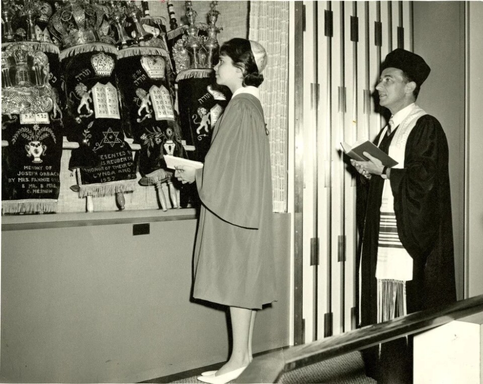 In a black and white photo, a young, white teenage girl stands in a Jewish synagogue facing a row of Torahs. She wears a gown and low heels. A rabbi stands behind her with an open prayer book. 