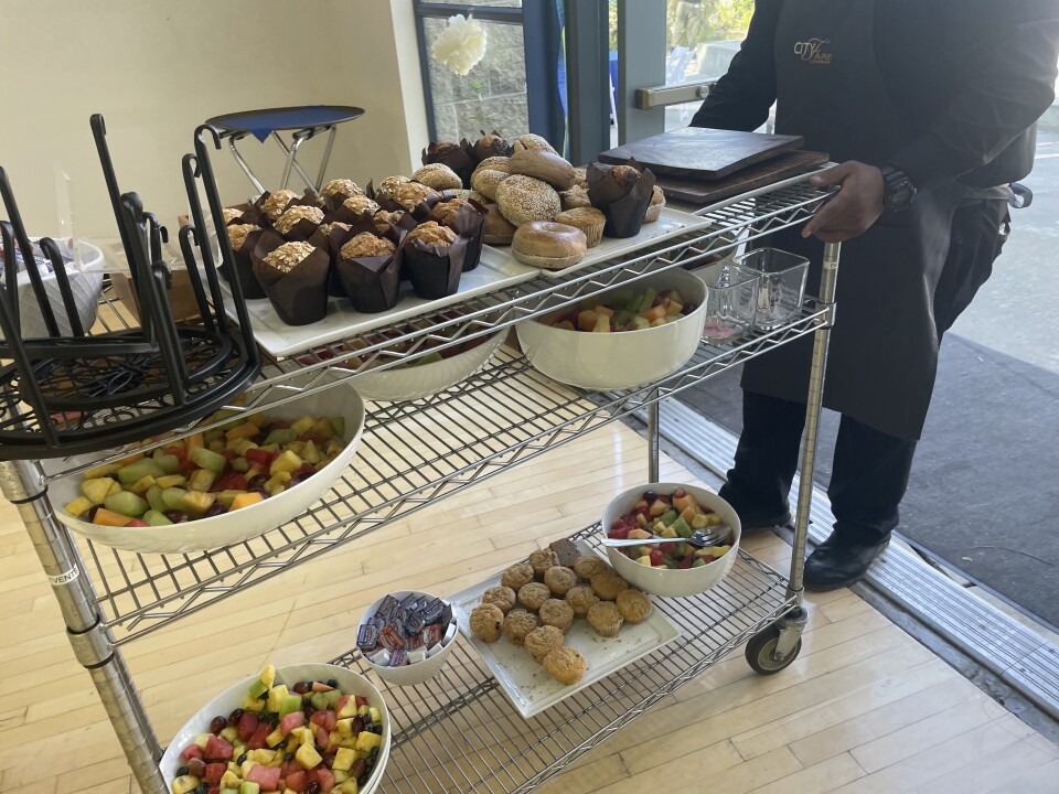 Person wearing an apron pulls a metal cart with muffins, bagels, and bowls of cut fruit. 