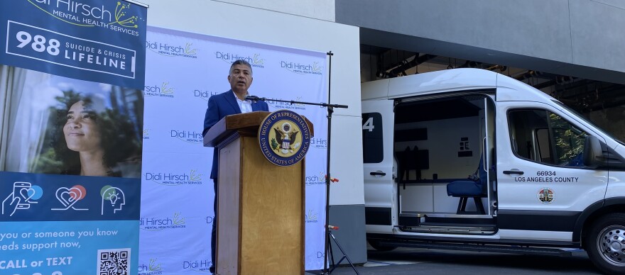 Congressman Tony Cárdenas stands at a podium. He wears a blue suit and to his right is a banner which has information for 988 -- the suicide and crisis lifeline. To his left is a white van that says 'Los Angeles County' on the door. It's one of L.A. County's therapeutic transport vans, which are staffed by unarmed teams who respond to mental health crises.