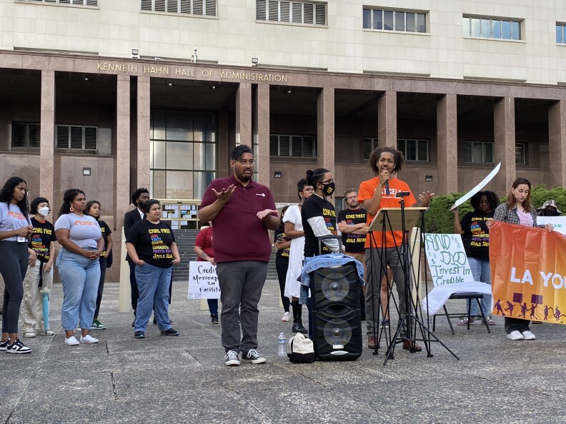 Dozens of protesters gathered in front of the Kenneth Hahn Hall of Administration to call on L.A. County Board of Supervisors to bring online 3,600 mental health treatment beds, among other demands. Mark-Anthony Clayton-Johnson stands holding a mic at the podium, speaking to the crowd. He wears a bright orange t-shirt that reads "Justice LA"