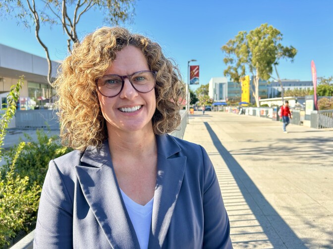 A woman with light-tone skin with glasses and wavy hair stands by a walkway during the day.