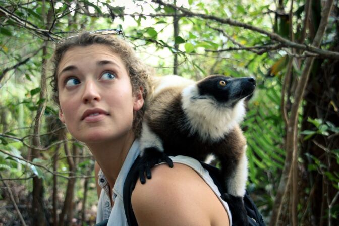 Alizé Carrère looks upward to her left as a small, furry, black, white and brown mammal rests on her shoulders.