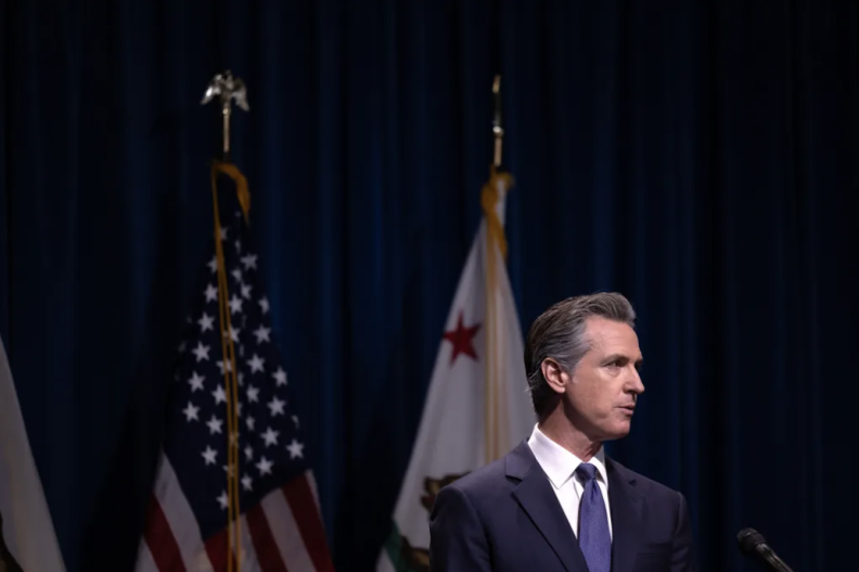 At the lower right of this image is a suited man with light skin tone speaking at a podium, the United States and California state flags behind him.
