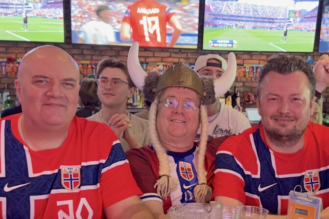 Three men with light skin tone wearing red jerseys with a navy blue cross sit in front of several TVs showing a soccer game. 