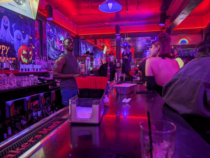 Interior of a bar with Halloween decorations. A black bartender is talking to a drag queen.