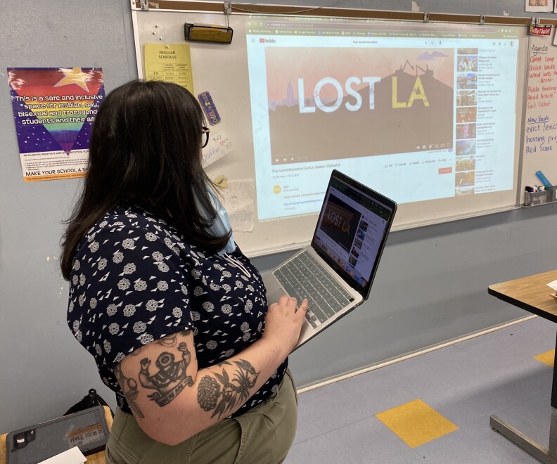 A woman stands facing a white board in a classroom. A projection on the white board mirrors the screen of a laptop she's holding in her hands as she stands.