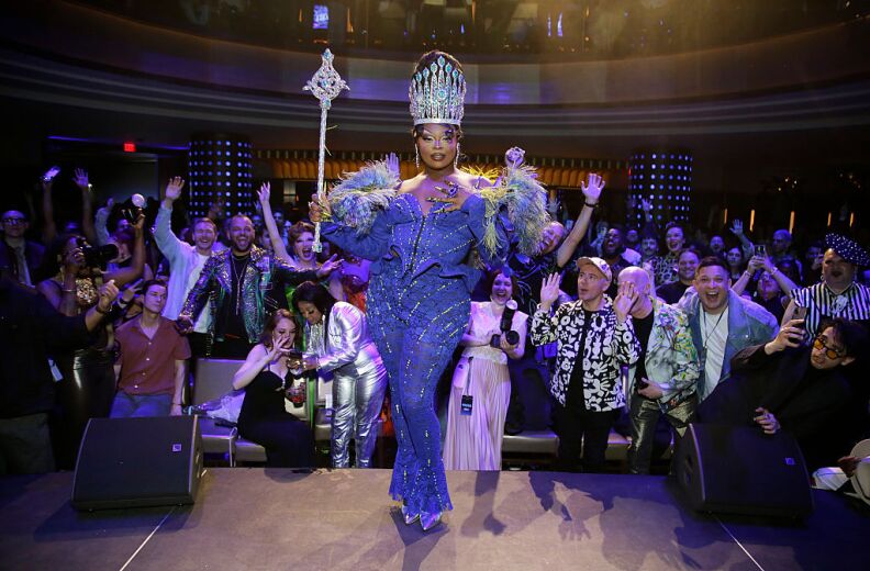 A drag performer dressed in immaculate blue outfit strikes a pose as an excited crowd cheers behind them.