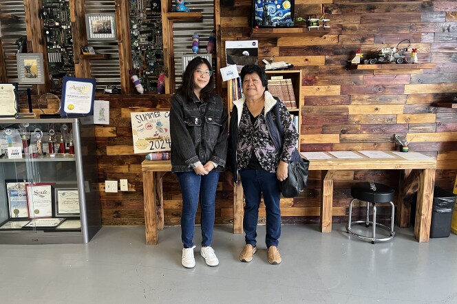 A mother and daughter with dark hair and medium skin stand in front of a wood-covered wall that's covered in award certificates and tech gadgets.