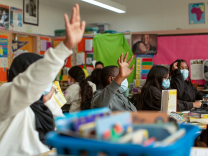 A group of children sitting at their desks raising their hands. There is a blurry blue bucket with school supplies at the front center of the photo. In the background are colorful papers hanging from the wall.