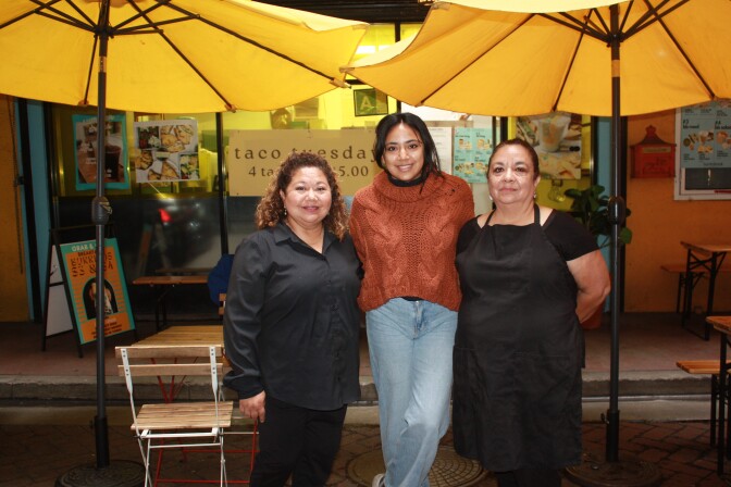 A group of three medium-skinned women stand outside of a cafe. Behind them are two large yellow umbrellas and a white chair. The woman with dark hair in the middle is wearing jeans and an orange sweater, and the women on both sides are dressed in black. 