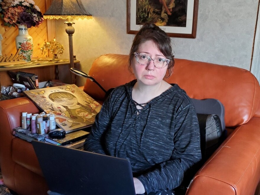 A 30 or 40-something white woman sits on a large leather chair with a laptop open on her lap. She looks mournfully at the camera.