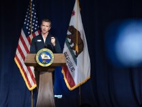 A man with light-toned skin stands at a lectern with the seal of the Governor of the State of California. He wears a blue zip-up jacket with a bear logo. Behind him are the U.S. and California flags. He's gesturing with his left hand.