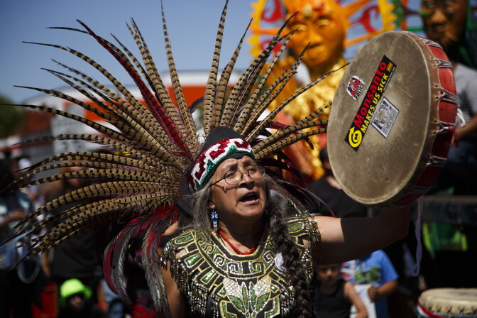 A man with medium-tone skin wear a feathered headdress and carroes a drum