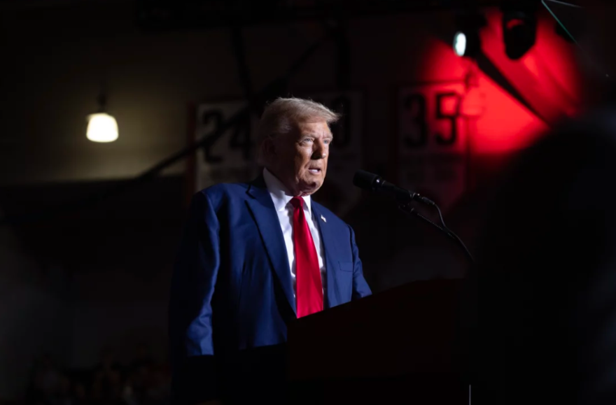 A man wearing a blue suit jacket and a red tie is standing behind a podium.