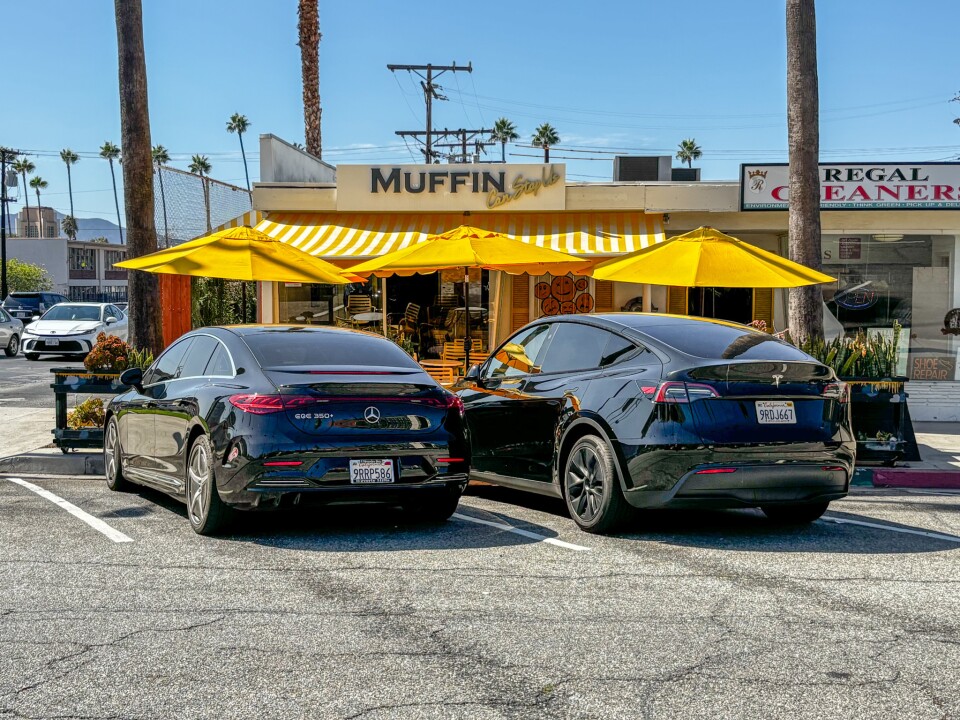 The storefront of Muffin Can Stop Us café, with yellow-and-white striped awnings and umbrellas shading outdoor seating. Two black luxury cars are parked in front.