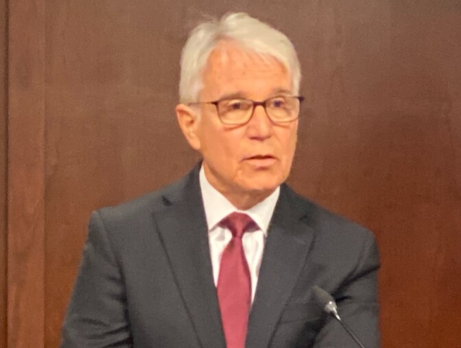 George Gascón, a light-skinned Latino man wearing a dark suit jacket with a red tie and white shirt, speaks at a podium.